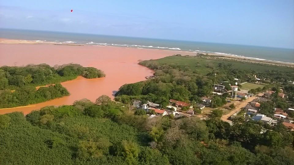 Vista aérea da foz do Rio Doce tomada pela lama alaranjada de rejeitos em novembro de 2015, com a vila de Regência ao lado e barreiras de contenção na água