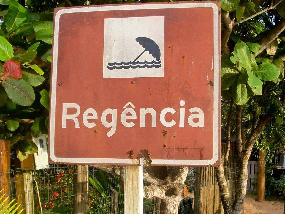 Placa marrom de sinalização turística com o nome Regência, enferrujada pelo tempo, entre a vegetação na entrada da vila