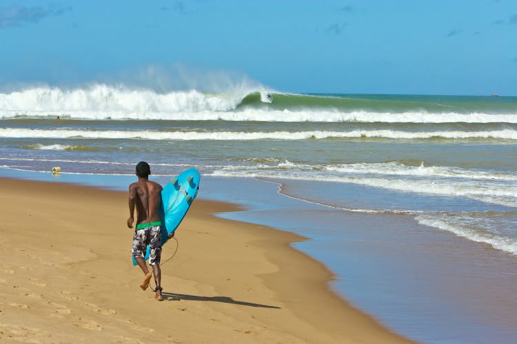 Surfista caminha pela areia de Regência com a prancha debaixo do braço, enquanto uma esquerda longa quebra ao fundo